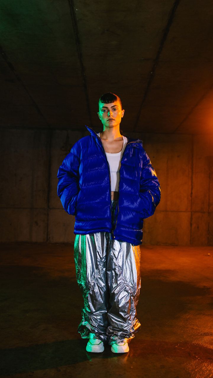 A woman in a cobalt puffer jacket and silver trousers in a car park at night